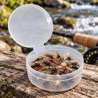 Open translucent hinged-lid storage cup holding fishing flies beside a stream